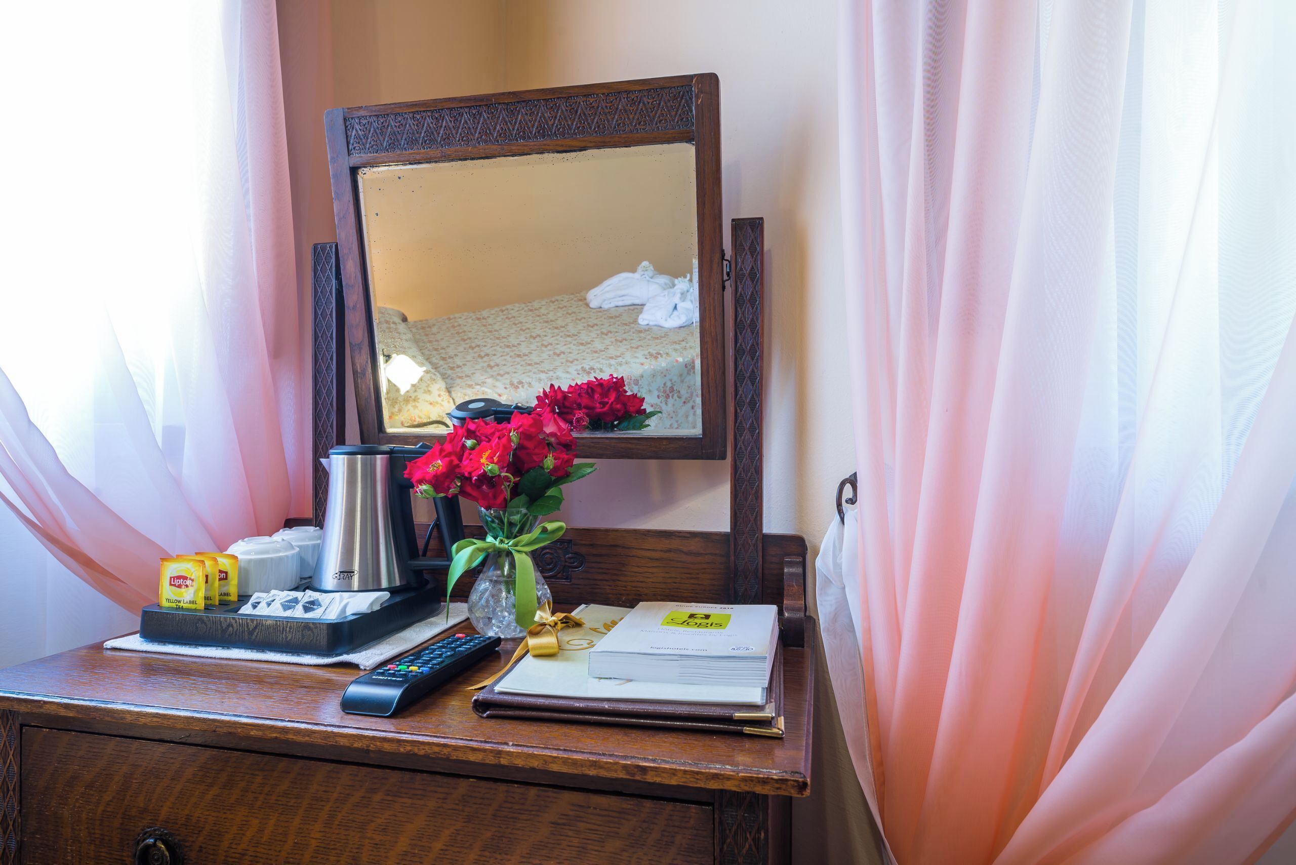 A wooden desk with a remote control, a coffee pot, a vase with flowers, and a book.