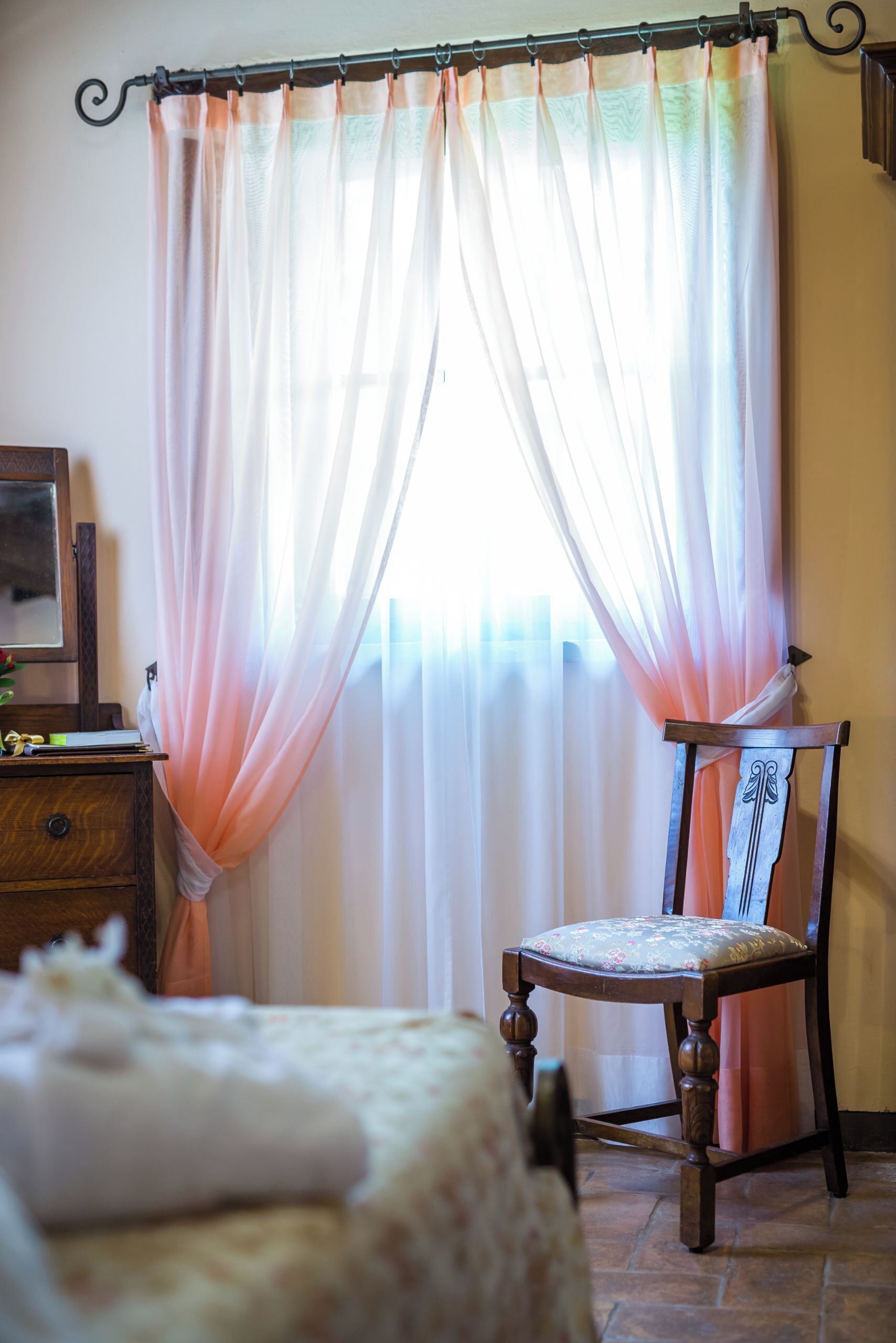 A room with a bed, a wooden chair, a nightstand, and sheer curtains.