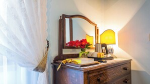 A wooden dresser with a mirror, a vase with red flowers, a lamp, and a book. - Deluxe Double Room (DERUTA)