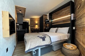 A compact bedroom with a bed, built-in storage, a small kitchenette, and a wooden accent wall. - Les Cerfs - Appartement & Studio (Ayent)