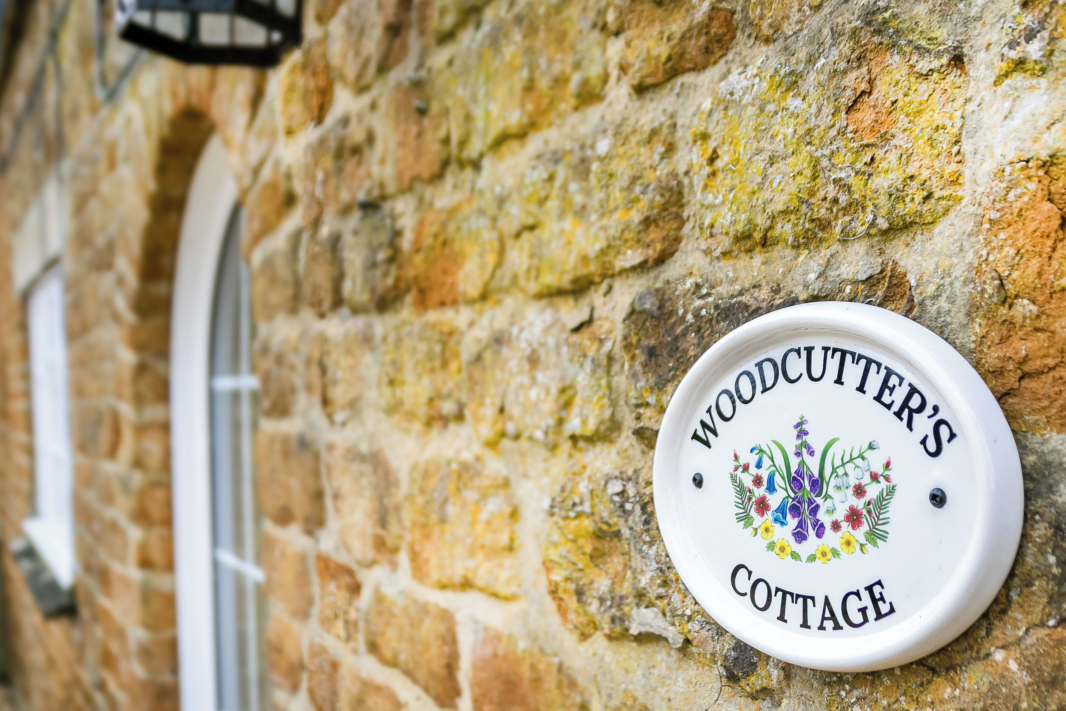 A white sign with "Woodcutters Cottage" and a floral emblem on a stone wall.