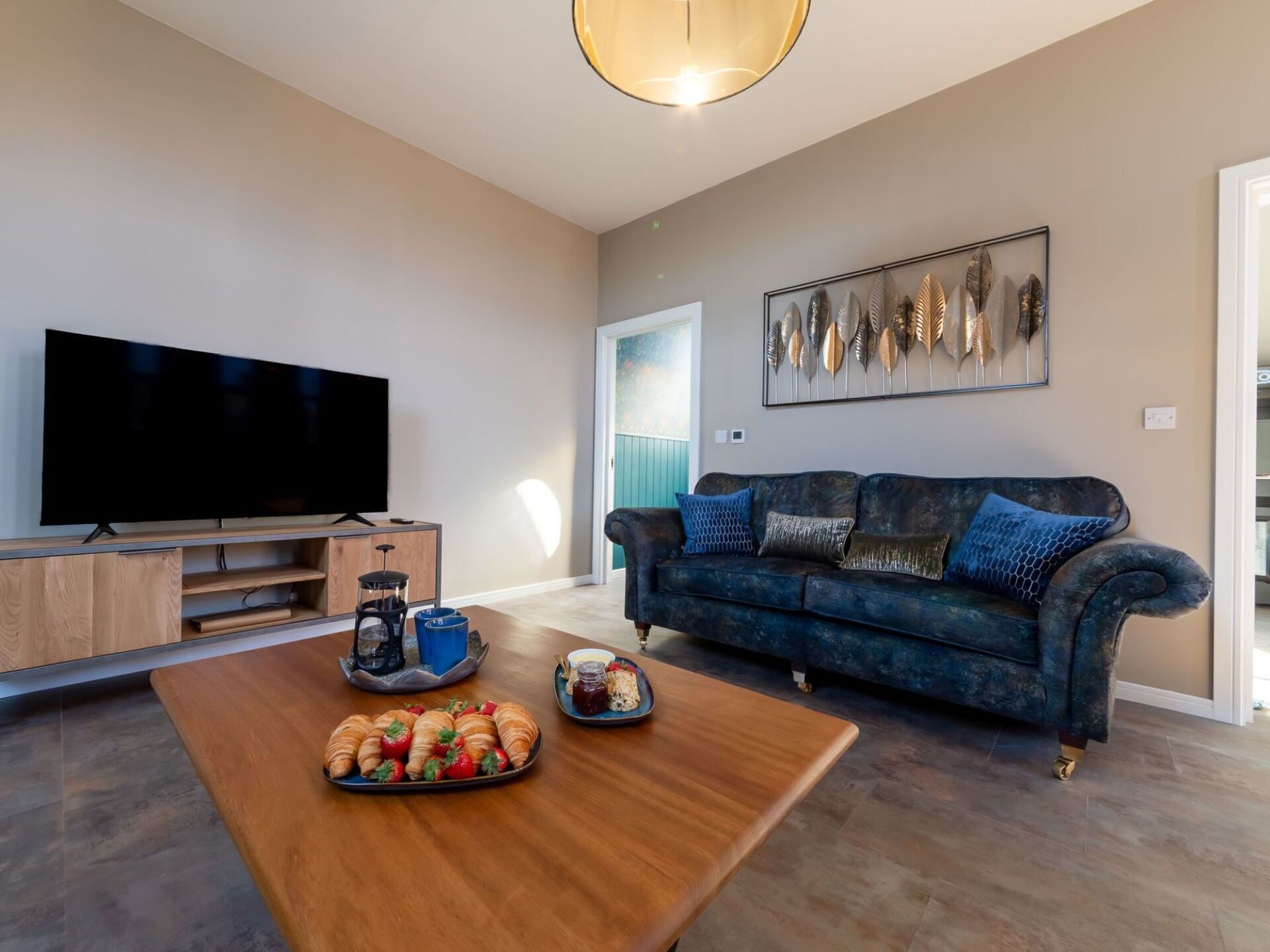 Un salon moderne avec un téléviseur à écran plat, une table basse en bois sur laquelle sont posés des croissants, un canapé bleu agrémenté de coussins à motifs et une œuvre d’art accrochée au mur.