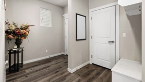 A hallway with a wooden floor, a white door, a framed picture on the wall, and a vase with flowers on a small table.