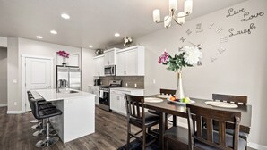 A modern kitchen with white cabinetry, a central island with bar stools, a dining area with a wooden table and chairs, and a wall clock with motivational phrases.