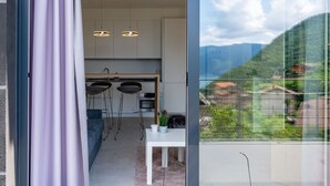 A modern kitchen with two pendant lights, a dining area with a white table and bar stools, and a view of a mountainous landscape through large windows. - Nice apartment for 3 people with WIFI, A/C, pool, TV and terrace (Nesso)