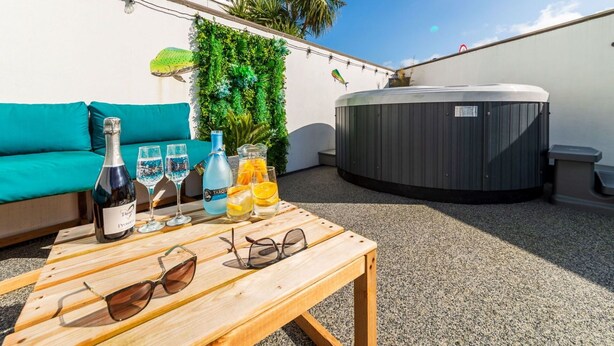 A rooftop terrace with a teal sofa, a wooden table with drinks and glasses, a hot tub, and a wall with greenery and a sign for a cinema.
