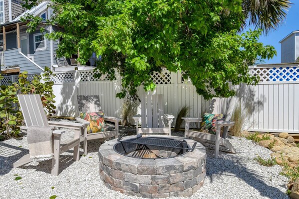 A backyard with a stone fire pit, wooden chairs, and a white fence.