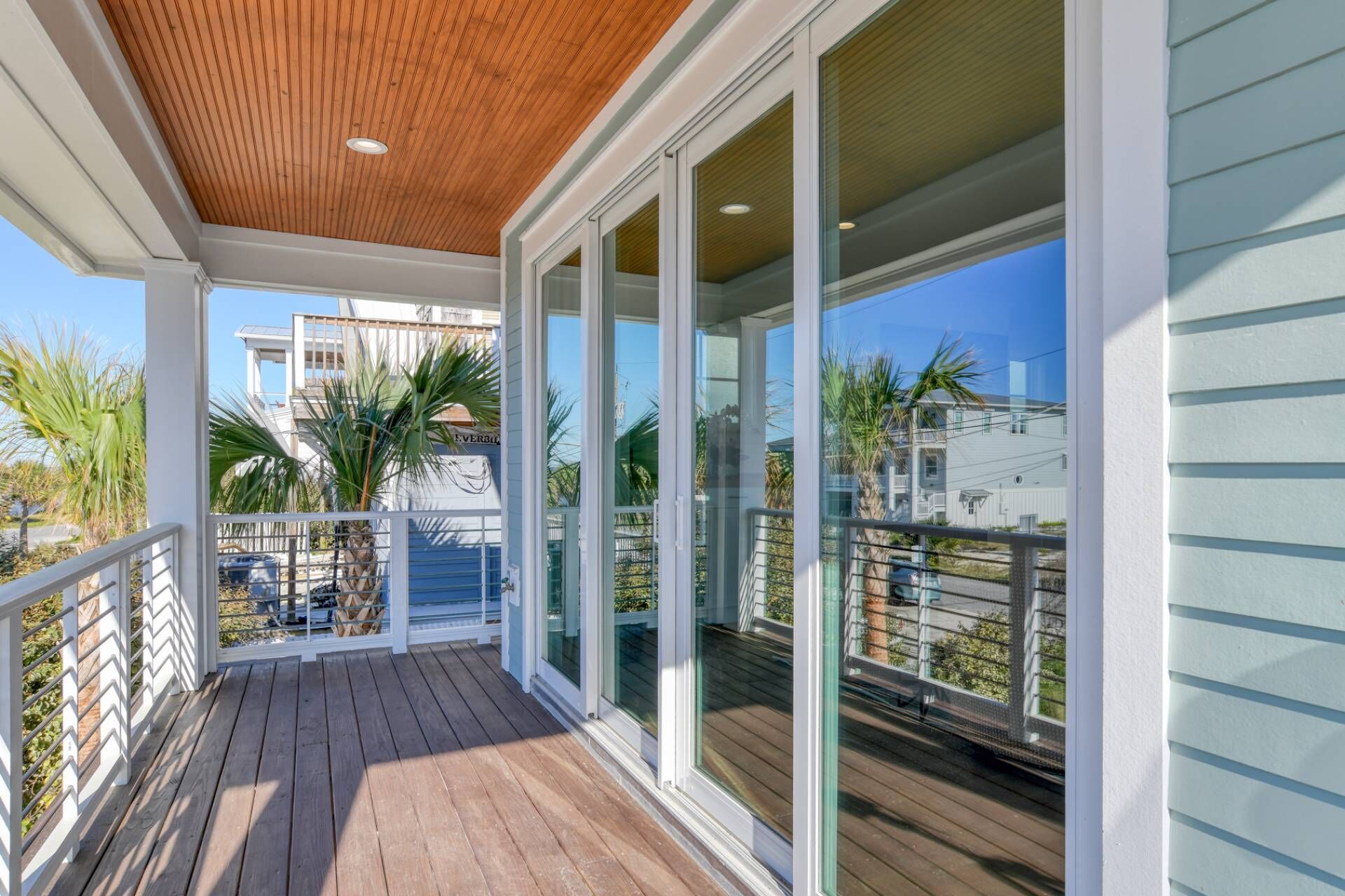 A covered wooden deck with glass doors leading to a balcony, overlooking a residential area with houses and palm trees.