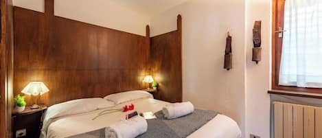 Quarto de hotel com cabeceira de madeira, duas luminárias de apoio, cama com roupa de cama branca, manta cinza e radiador.
