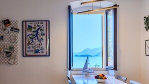 Uma área de refeições com uma mesa, cadeiras, uma tigela de frutas e uma taça de vinho. Um pôster de Lago Di Como está na parede.