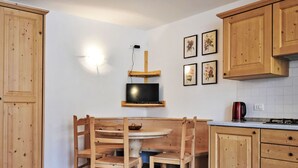 Keittiössä on puiset kaapit, ruokapöytä tuoleineen, seinälle asennettu televisio ja vedenkeitin työtasolla.