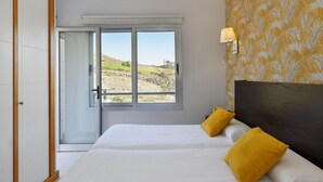 A bedroom with a large window, a bed with white bedding and two yellow pillows, and a wall with a yellow floral pattern.