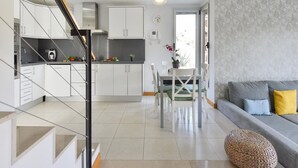 A modern kitchen with white cabinets, a dining area with a table and chairs, and a living area with a sofa and cushions.