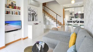A modern living room with a flat-screen TV, a grey sofa, a coffee table, and a staircase leading to a kitchen area.