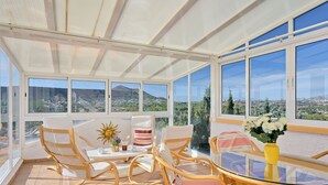 Une véranda meublée de mobilier en osier, dotée d’une table en verre et de grandes fenêtres offrant une vue panoramique.