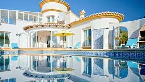 En pool med en klar vattenyta som speglar omgivningen, en täckt uteplats och en byggnad med en vit fasad och blå kakeldetaljer.