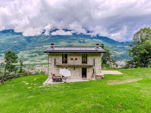 A stone house with a balcony, a table, and chairs outside, surrounded by a grassy area and mountains in the background. - Stunning private villa for 5 guests with WIFI, TV, balcony and pets allowed (Cosio Valtellino)