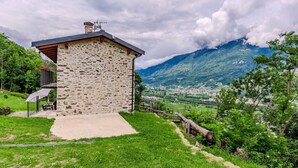 Una casa de piedra con balcón, rodeada de abundante vegetación y montañas al fondo.