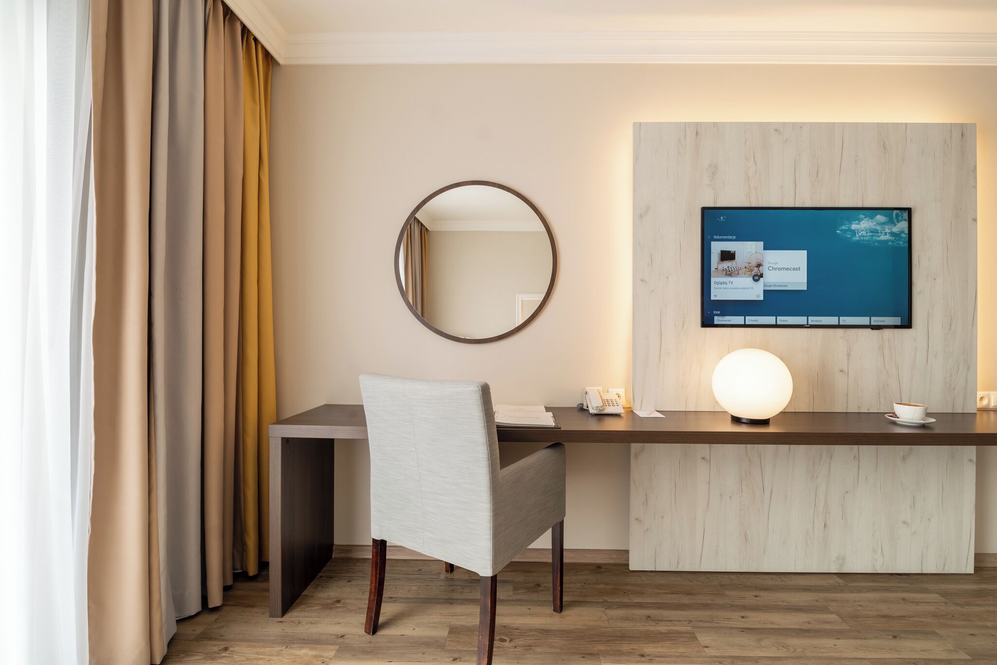 A hotel room with a desk, chair, mirror, and a television mounted on the wall.