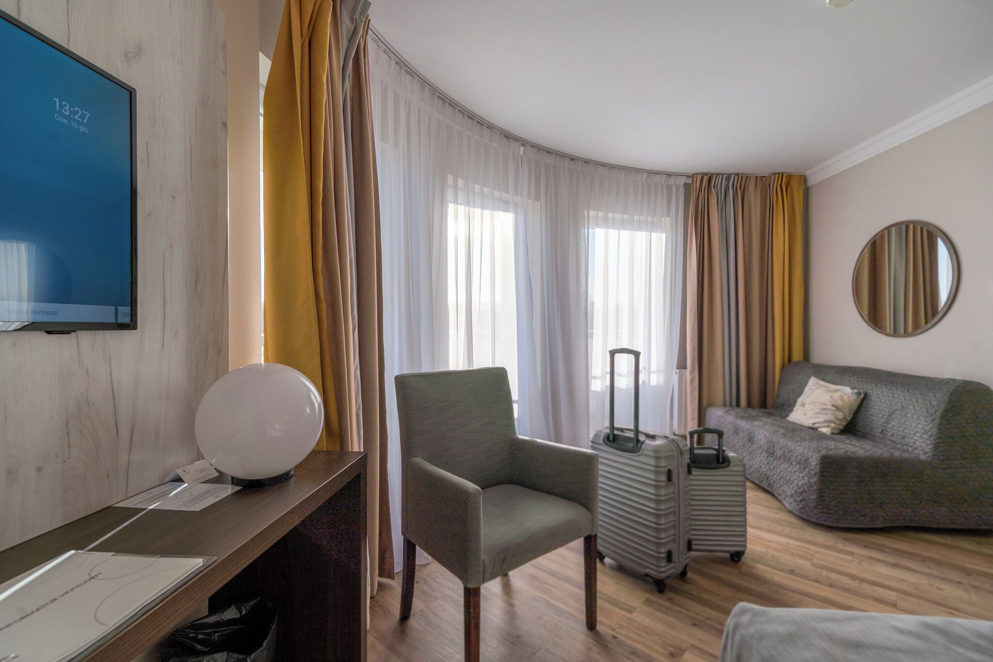 Camera d'albergo con una scrivania, una sedia, un divano, un tavolino, una lampada, una valigia e uno specchio a parete.