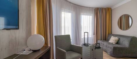 Una habitación de hotel con escritorio, una silla, sofá, una mesita, lámpara, maleta y espejo en la pared.