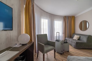 Una habitación de hotel con escritorio, una silla, sofá, una mesita, lámpara, maleta y espejo en la pared.