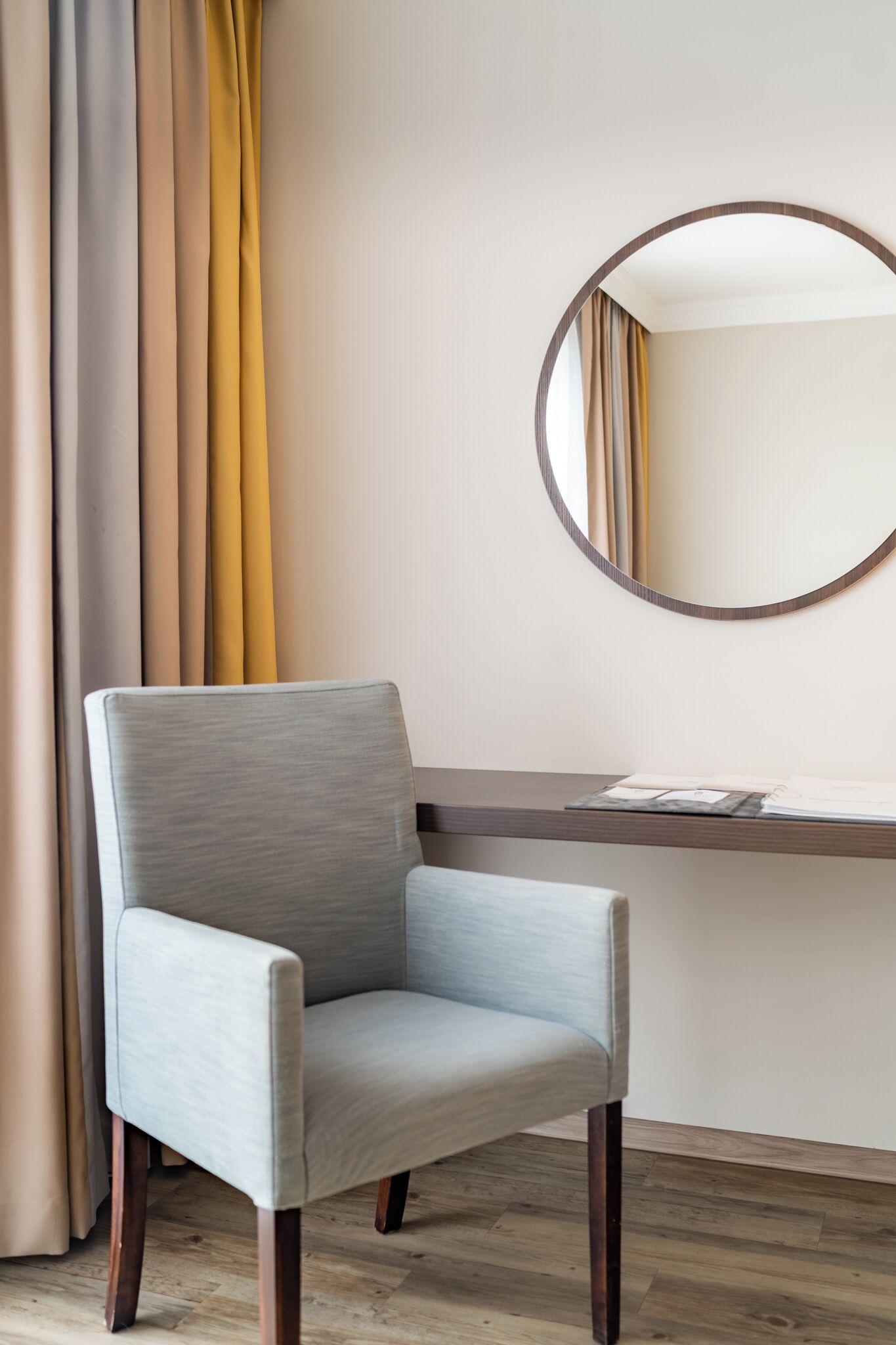 A chair with a light fabric, a wooden desk with papers, and a round mirror on the wall.