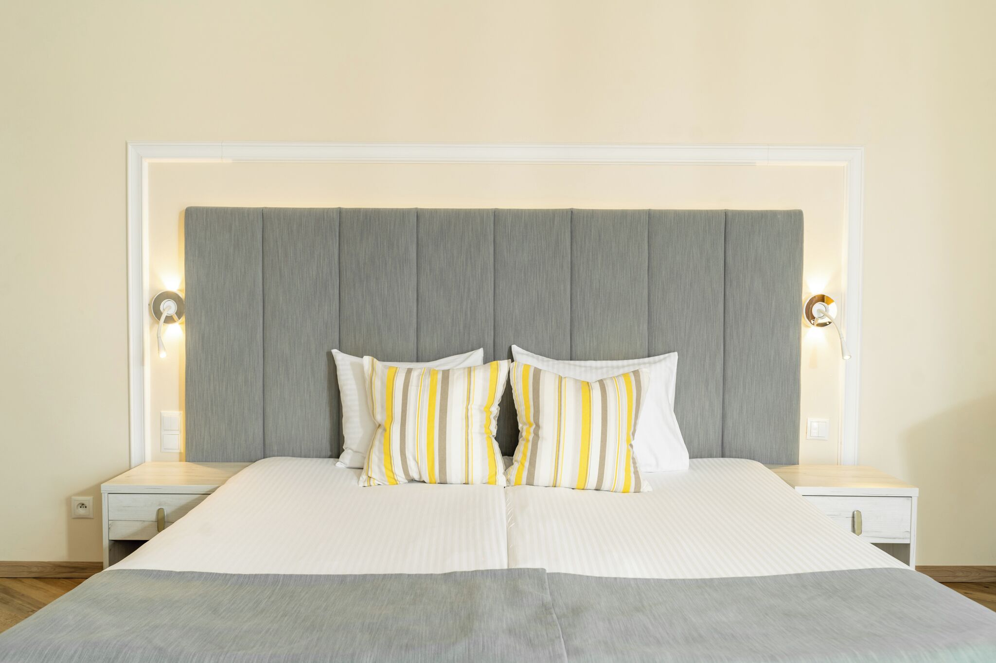 A neatly made bed with a grey headboard, two bedside tables with lamps, and a white wall.