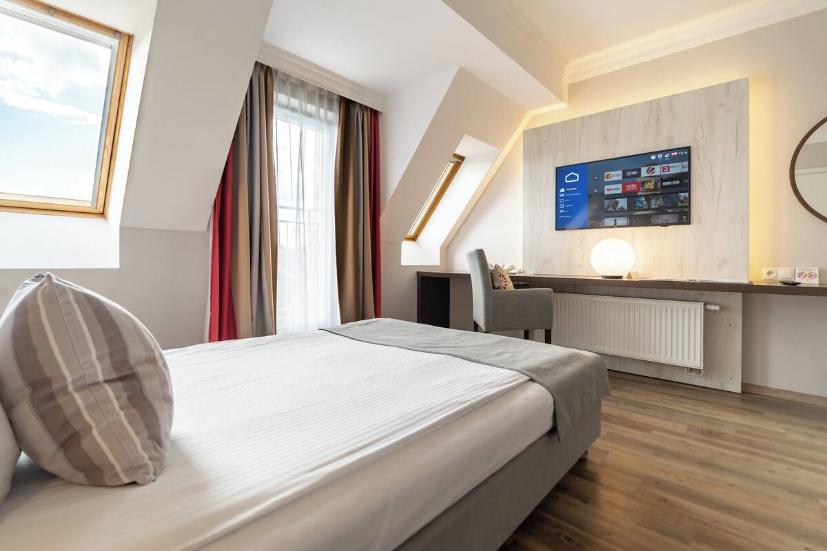 Une chambre d’hôtel moderne équipée d’un lit, d’un bureau, d’une chaise, d’un téléviseur fixé au mur et d’une fenêtre avec des rideaux.