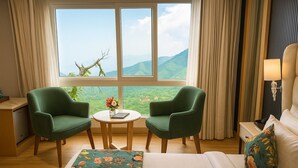 A hotel room with a bed, two green armchairs, a small round table with a vase of flowers, and a view of green hills through large windows. - Hotel Rockland Residency (Dehradun)