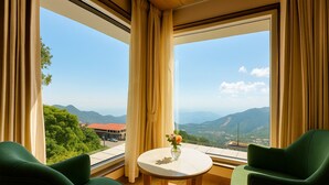 A room with a large window offering a mountain view, two green chairs, and a small table with a vase of flowers. - Hotel Rockland Residency (Dehradun)