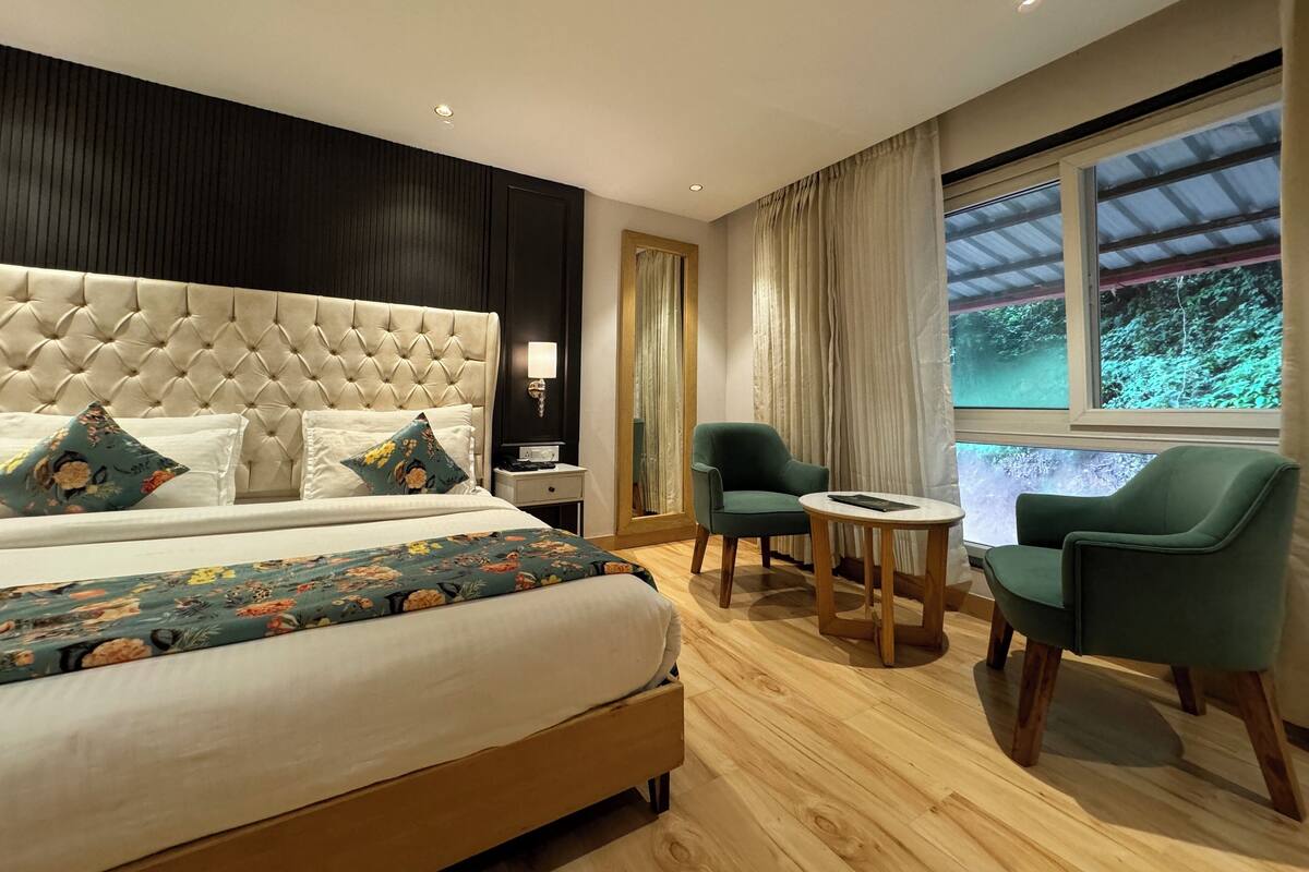 Un dormitorio con cabecera capitonada, cama con ropa de cama floral, dos sillas, una mesita y una ventana con vistas a zonas verdes.