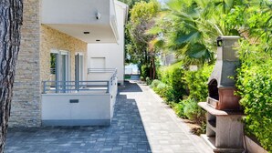 A modern building with a stone facade, a balcony, and a paved pathway leading to a garden area with palm trees and a stone structure. - Nice apartment for 6 people with A/C, WIFI, TV and terrace (Zadar)