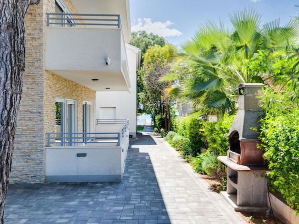 A modern building with a stone facade, a balcony, and a paved pathway leading to a garden area with palm trees and a stone structure.