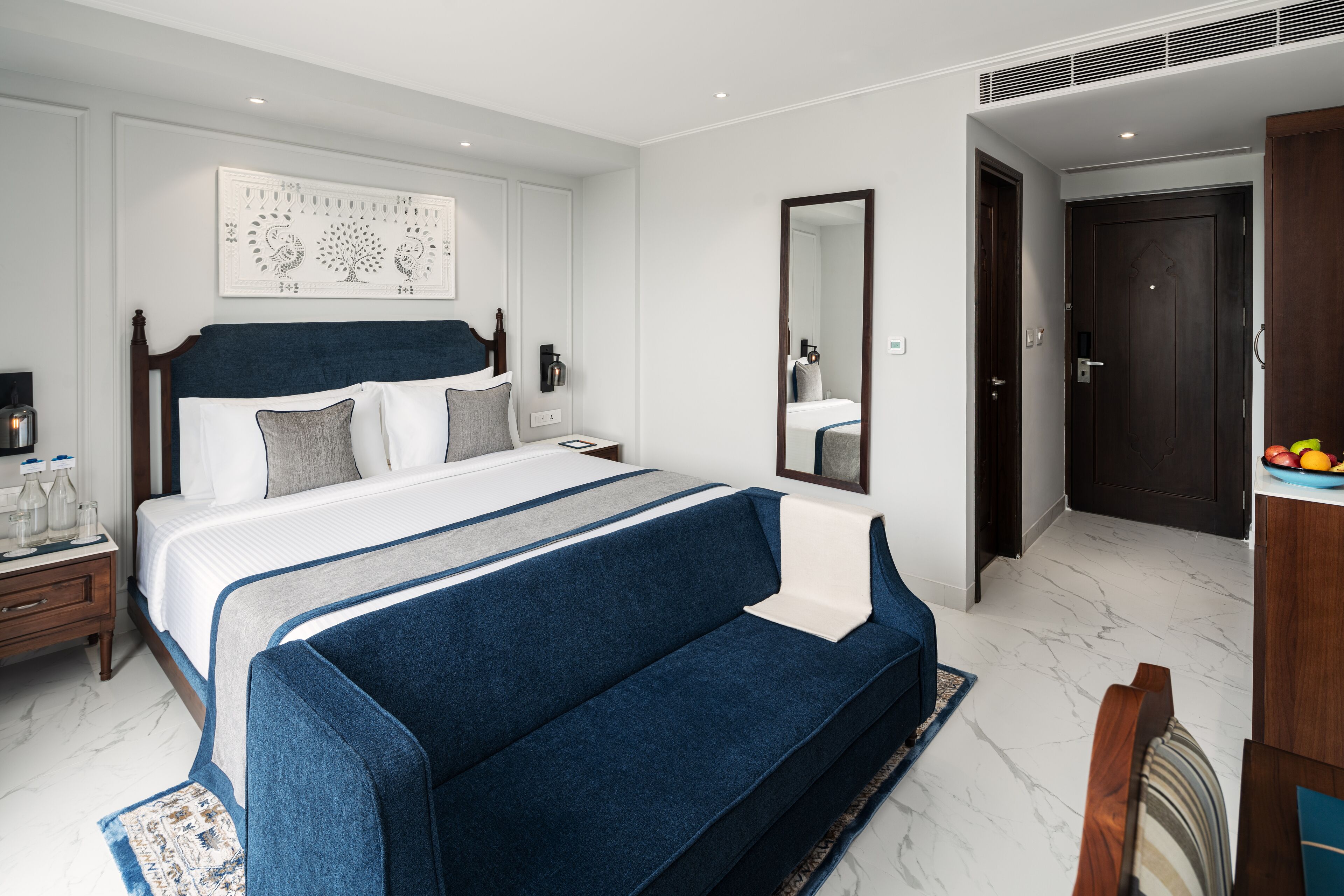 A modern hotel room with a large bed, a sofa, a bedside table with a lamp, a mirror, and a door leading to the bathroom.