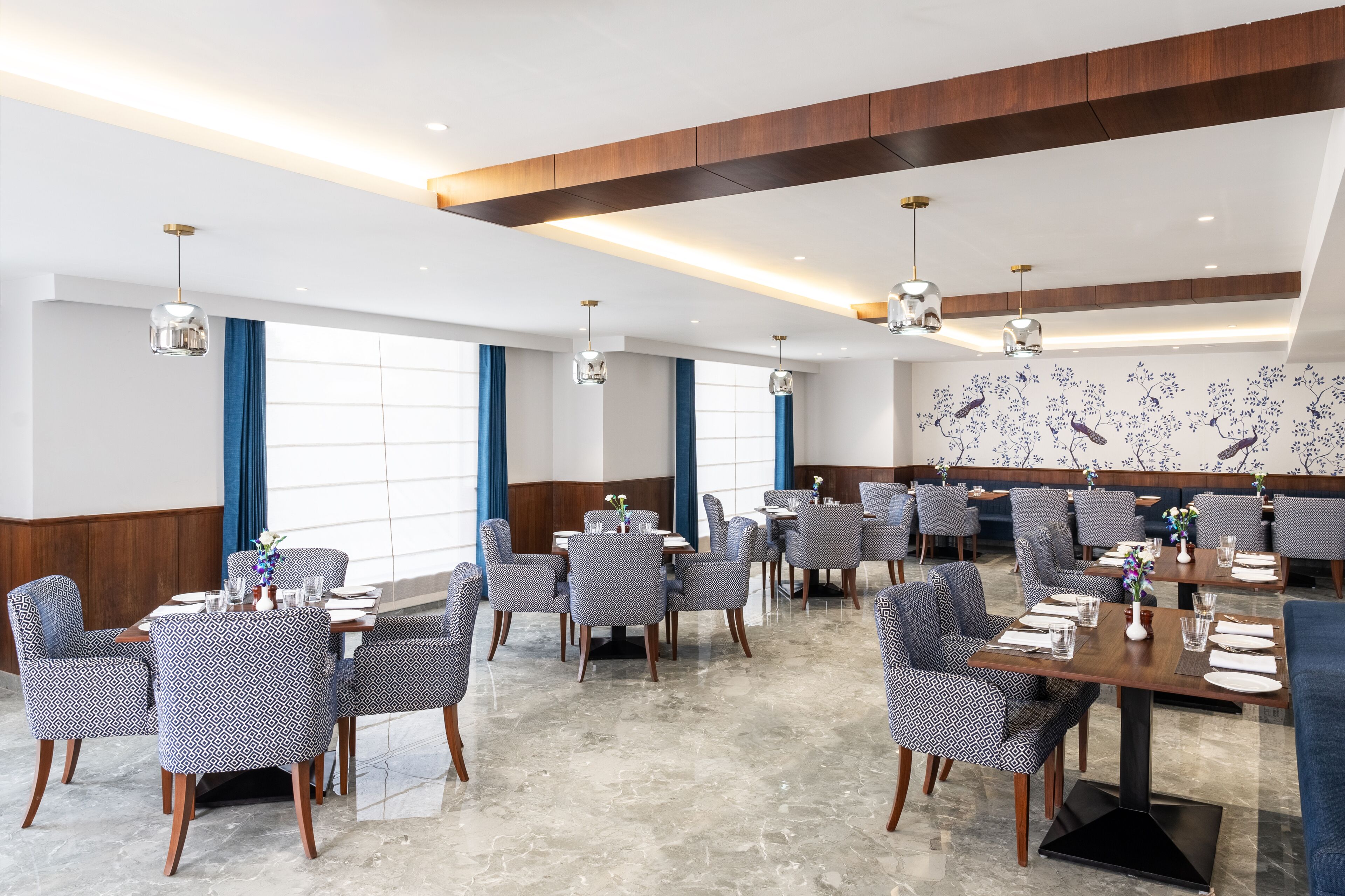 A modern dining area with high tables and patterned chairs, large windows with blue curtains, and a decorative wall mural.
