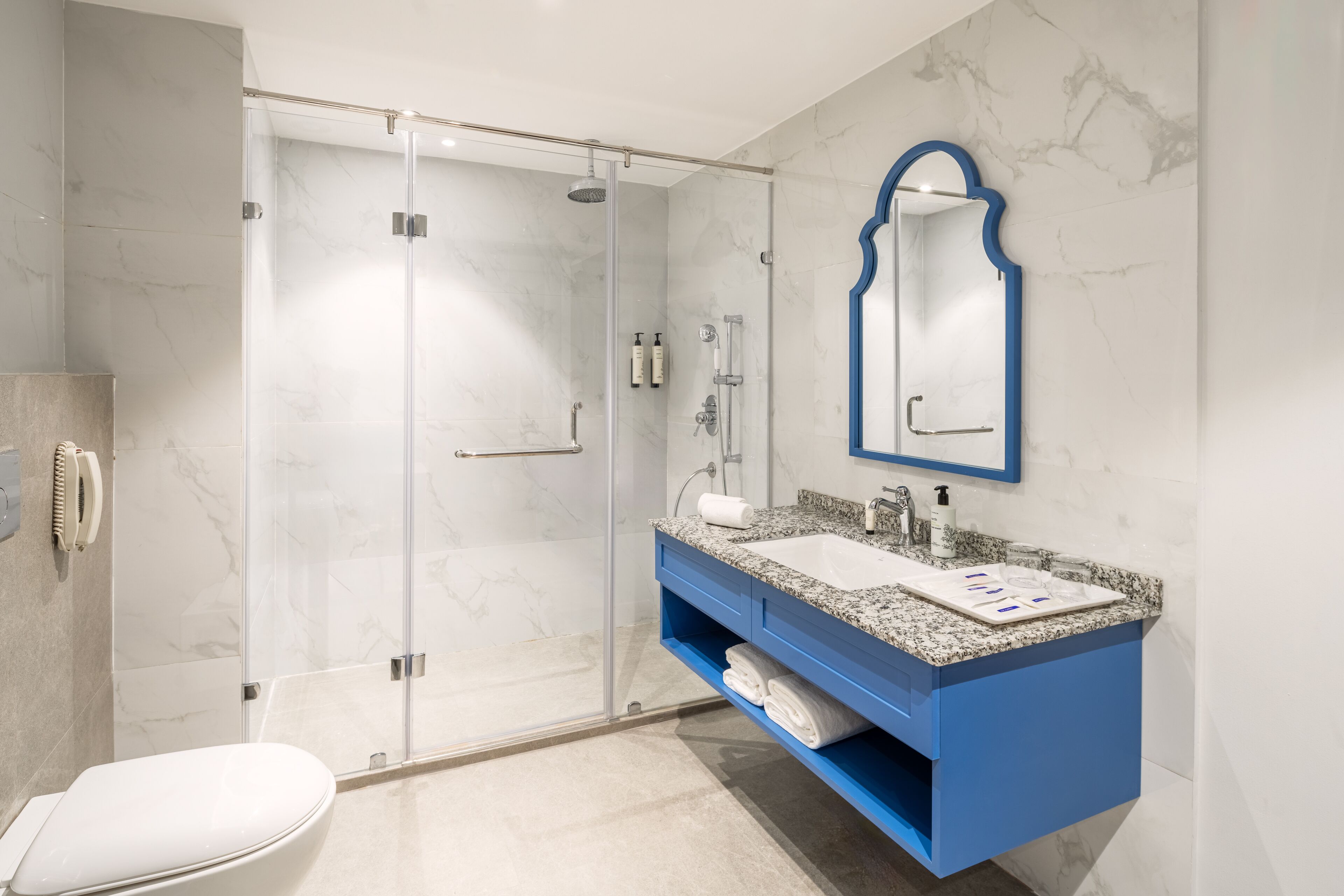 Een moderne badkamer met een douchecabine van glas, een wit toilet, een blauwe wastafel met een marmeren blad en een spiegel aan de muur.
