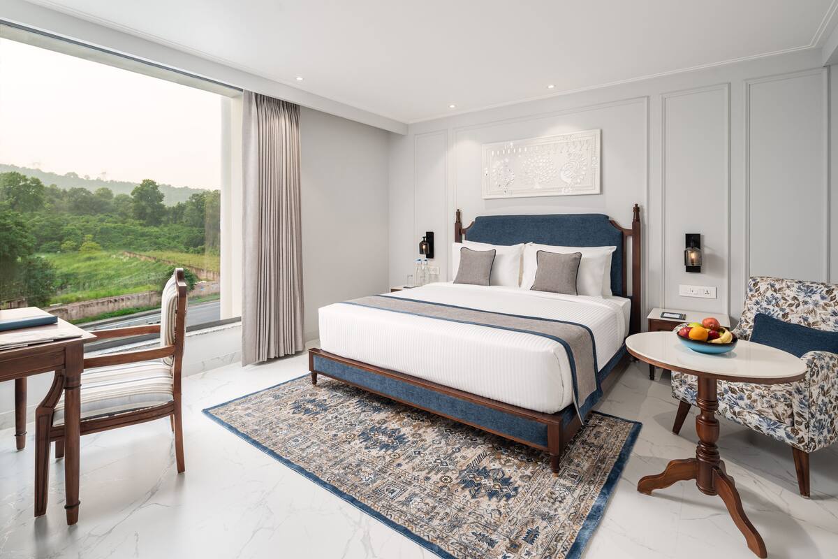 A modern hotel room with a large bed, a dining table, a chair, and a view of the outdoors.