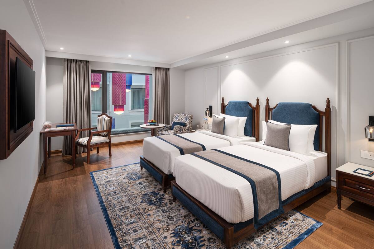 A hotel room with two beds, a TV, a desk, and a view of a building with colorful windows.