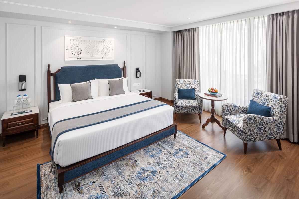 A hotel room with a large bed, two armchairs, a small table with a fruit bowl, and a decorative rug.