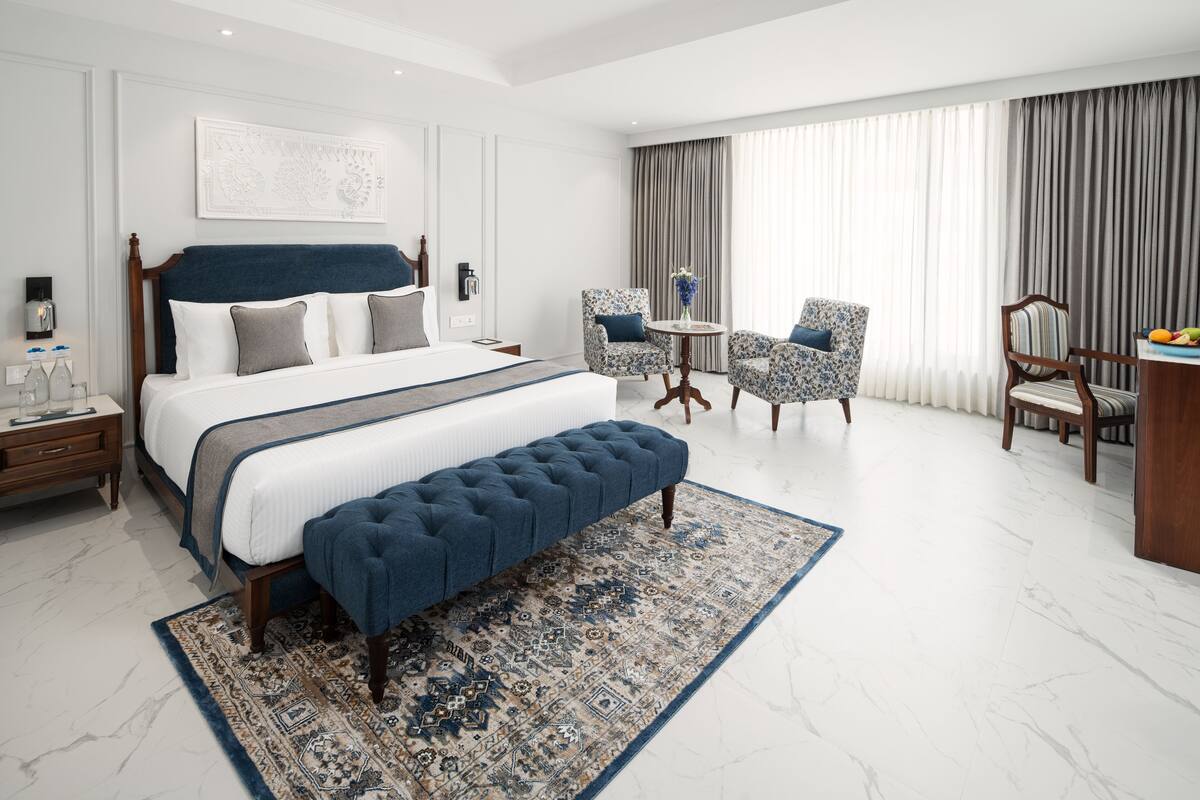 A hotel room with a large bed, a bench, two armchairs, a small table, and a desk with fruit.