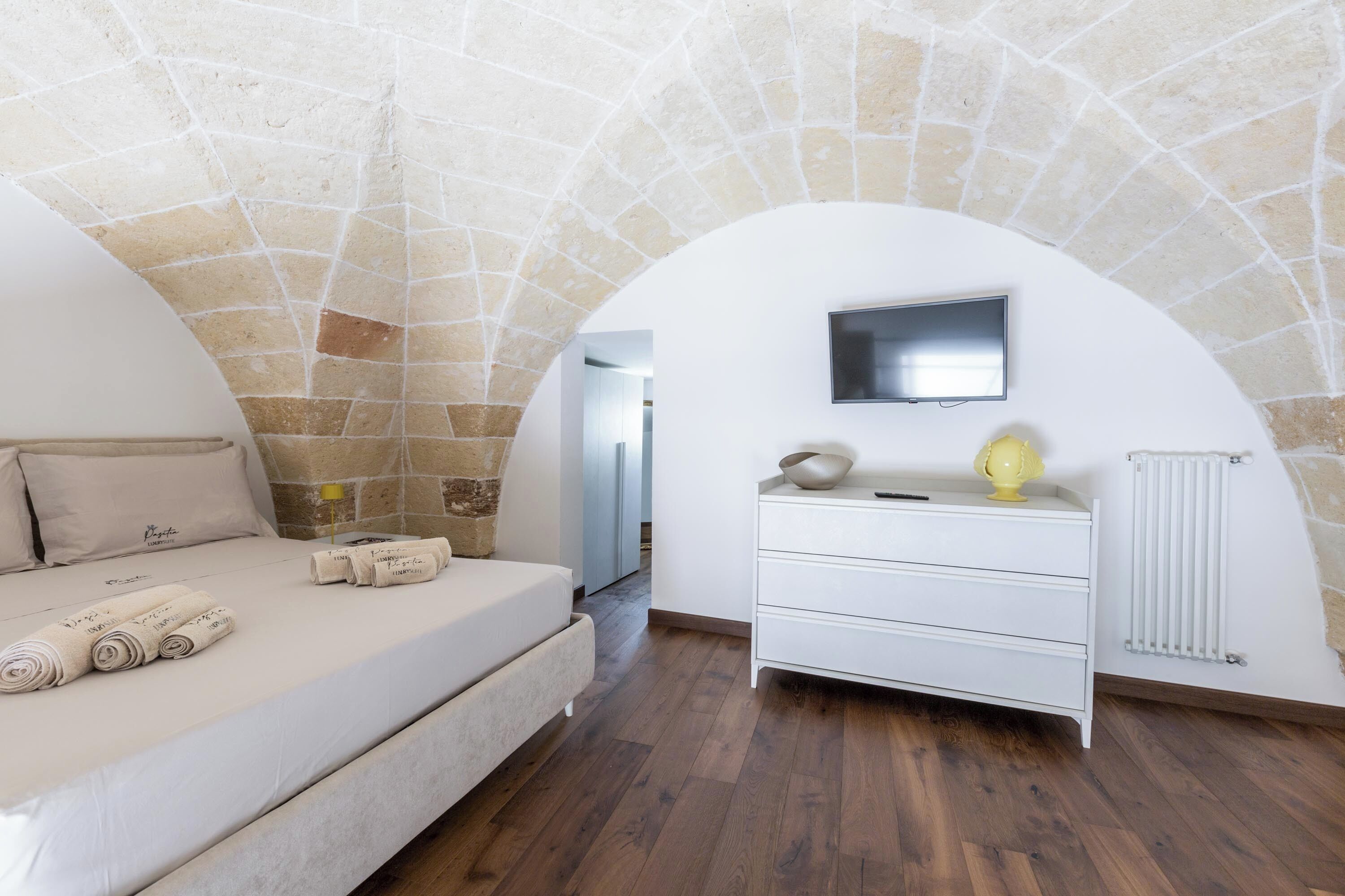 A bedroom with a stone ceiling, a bed with rolled towels, a white dresser, and a flat-screen TV.