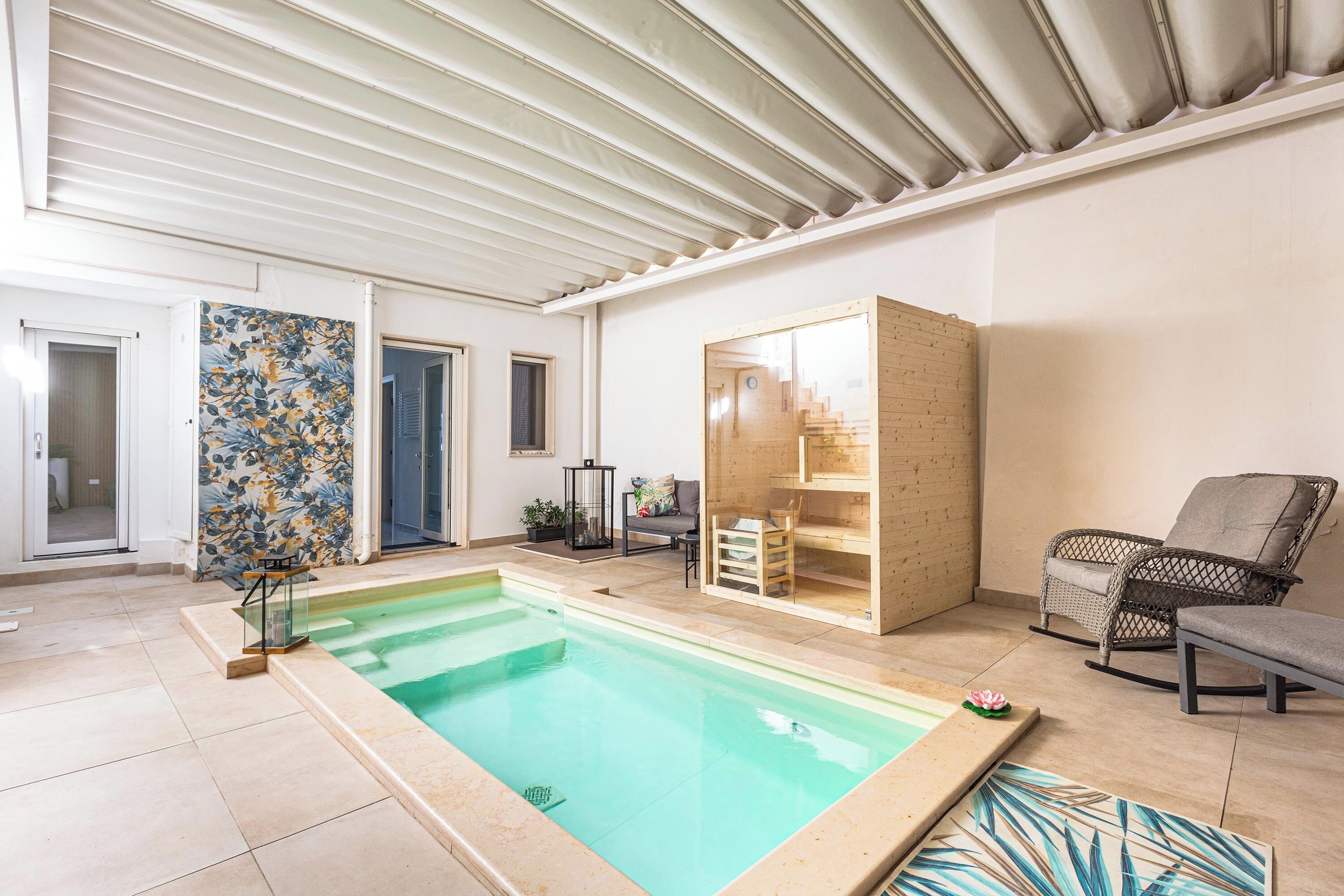 An indoor swimming pool with a glass enclosure, a wooden bench, a wicker chair, and a staircase leading to an upper level.