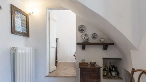 Ein schmaler Flur mit einer weißen Heizung, einem Holzstuhl, einem kleinen Tisch mit einer Schüssel und einem Regal mit Tellern und Flaschen.