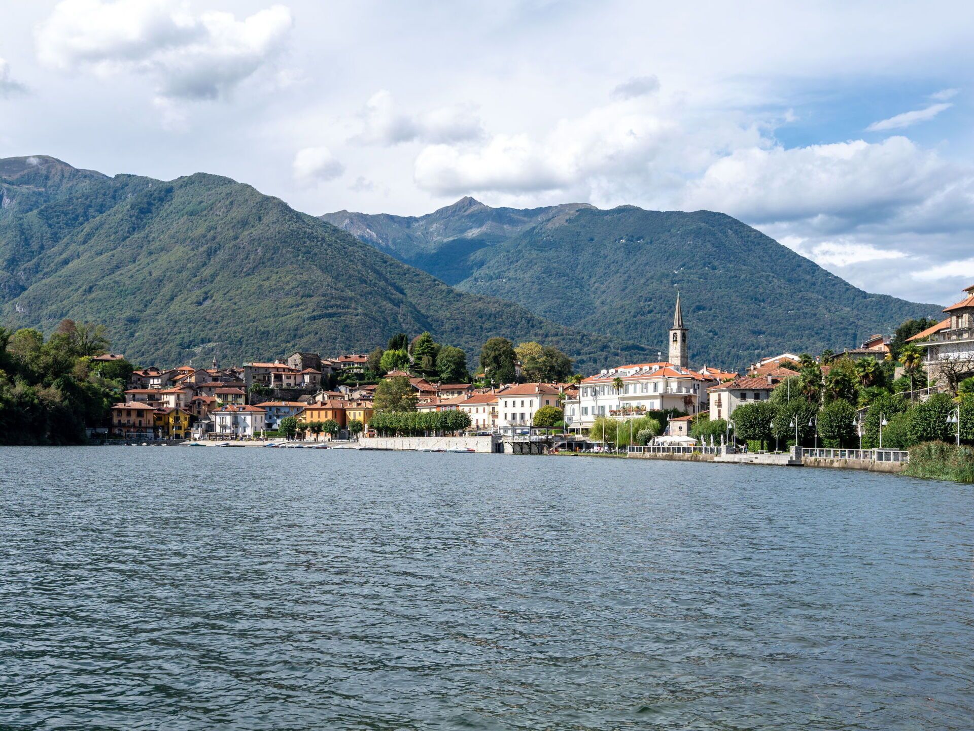 Un pueblo a orillas del lago con edificios a lo largo de la orilla, una destacada torre de iglesia y montañas al fondo.