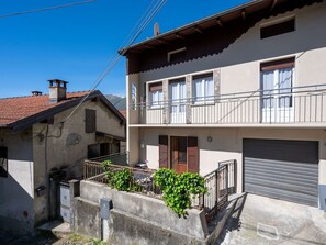 A two-story building with a balcony, a garage, and a small garden area with plants. - Beautiful  villa for 4 people with A/C, WIFI, TV, terrace and pets allowed (Mergozzo)