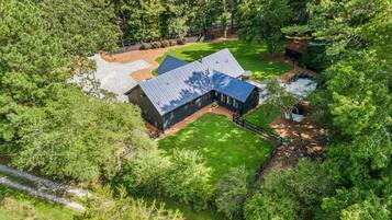 A large, dark-colored house with a sloped roof, surrounded by dense greenery and a small stream.