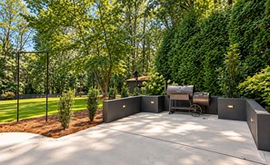 A modern outdoor patio with a built-in grill and seating area, surrounded by lush greenery and a well-maintained lawn.
