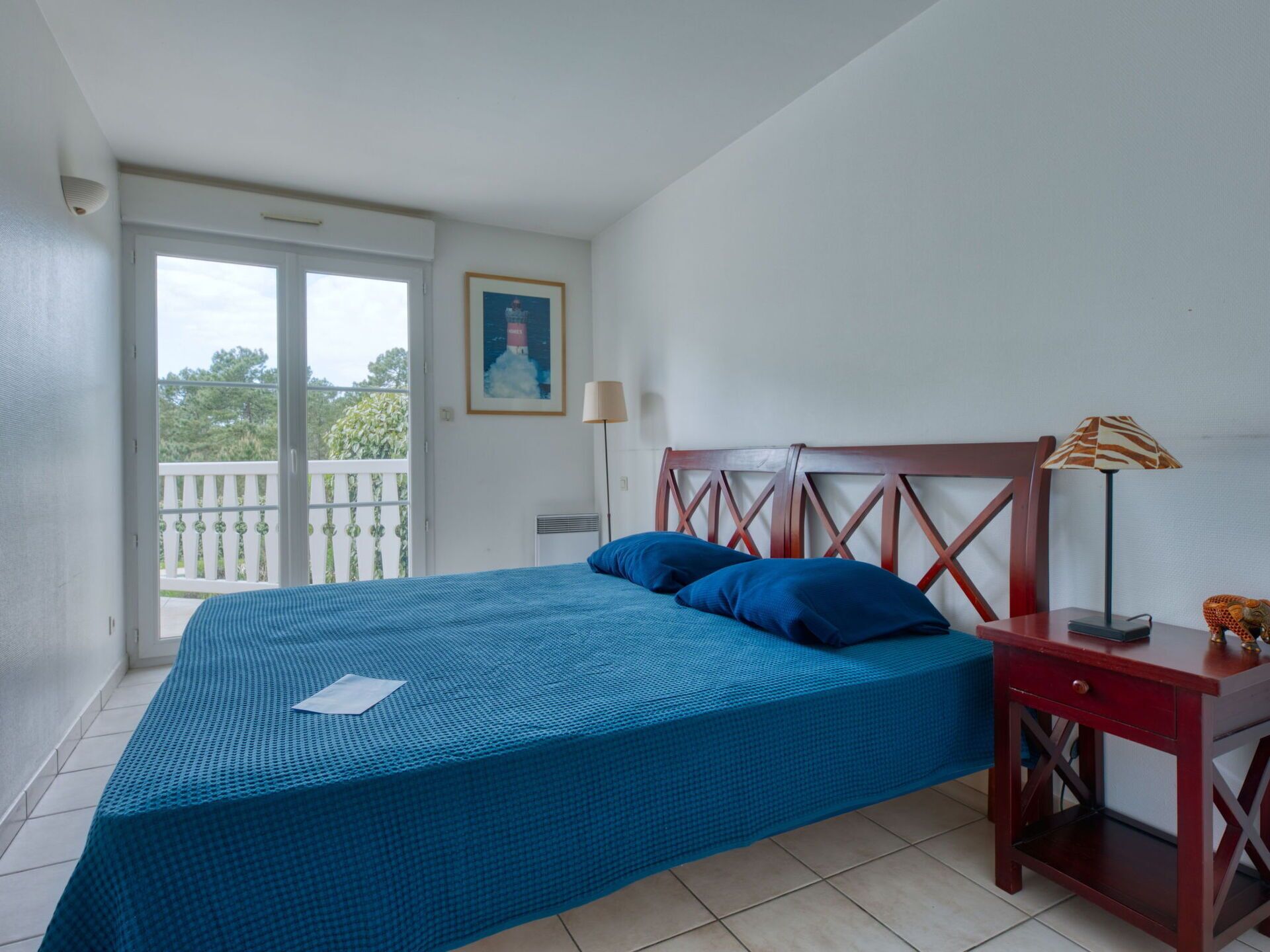Quarto com cama, mesa de cabeceira, abajur, quadro na parede e varanda com vista para árvores.