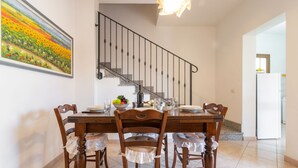 Un coin repas est aménagé avec une table en bois, dressée pour quatre personnes, et entourée de chaises dont les assises sont ornées de housses à volants. Un tableau représentant un champ de tournesols est accroché au mur, et un escalier avec une rampe en métal est visible au fond.